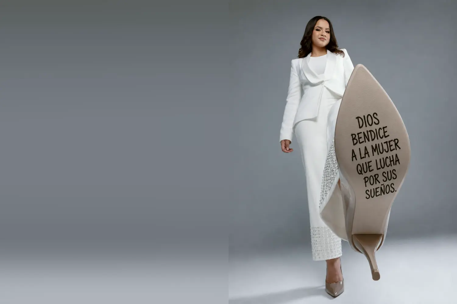 Yira Vargas camina con traje blanco junto a un zapato de tacón gigante en el que se lee «Dios bendice a la mujer que lucha por sus sueños».