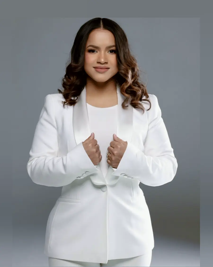 Retrato de Yira Vargas de pie, con traje blanco sobre fondo gris de estudio, mirando a cámara.